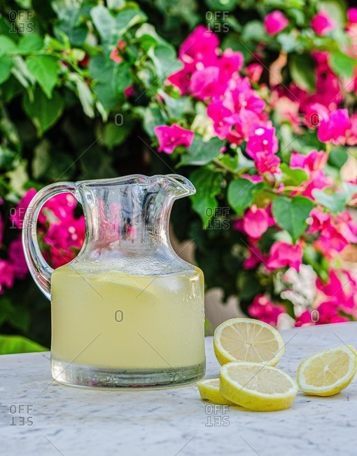 Glass jar with fresh cold lemonade placed on marble table with slices of lemon in summer garden with blooming plants in background