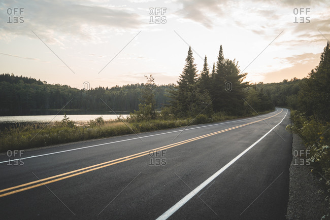 Asphalt road going near peaceful lake and green forest against cloudy sunset sky in La Mauricie National Park in Quebec, Canada