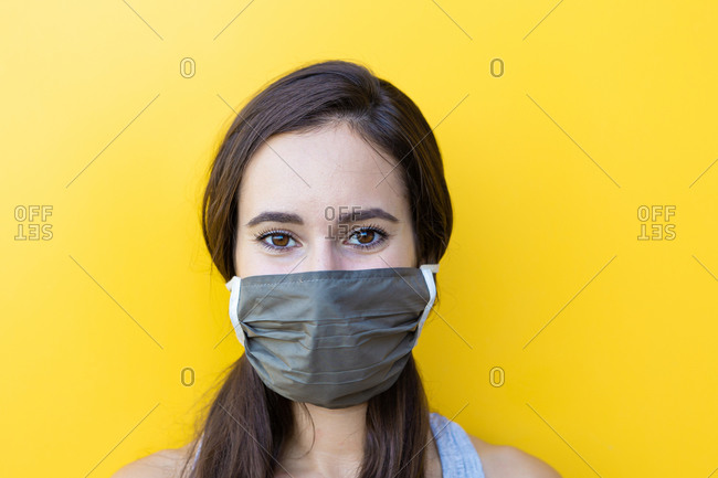 Isolated portrait of a beautiful young woman wearing a gray face mask  in front of a yellow wall