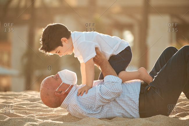 Grandfather with face mask during coronavirus outbreak plays with grandson at the beach