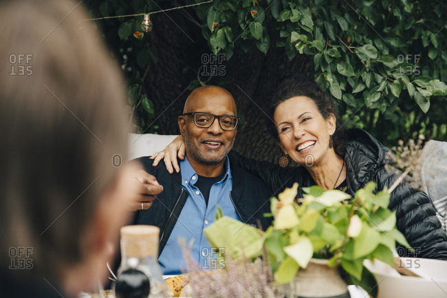 Cheerful senior woman sitting with arm around man while enjoying dinner party at back yard