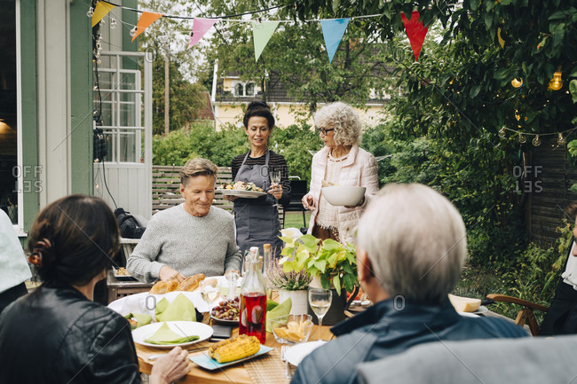 Active senior women bringing meal to friends sitting at dining table during garden party