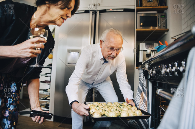 Senior woman standing by male friend putting tray of potatoes in oven at home