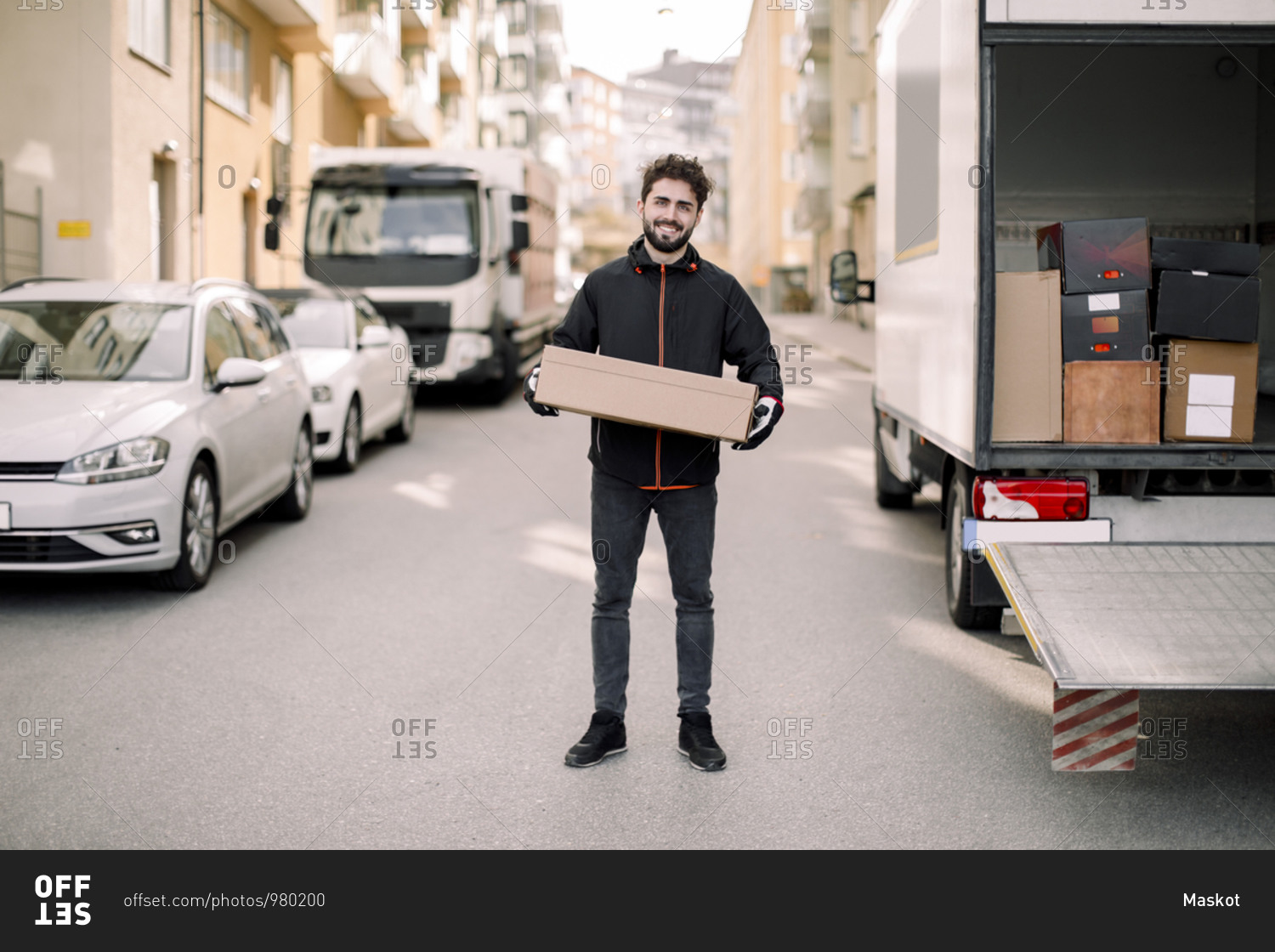 Portrait of confident young delivery man with box leaning on truck