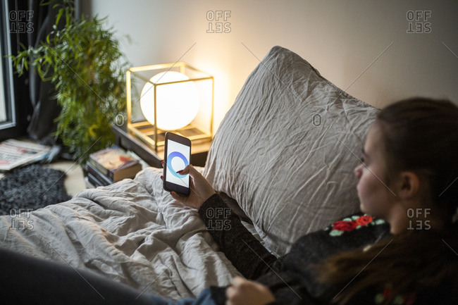 Teenage girl adjusting lighting equipment through smart phone app while lying in bedroom at smart home