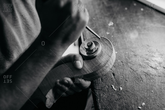 Black and white high angle of faceless craftsman carving swirl of violin head while working with wooden detail using bit at table in workshop