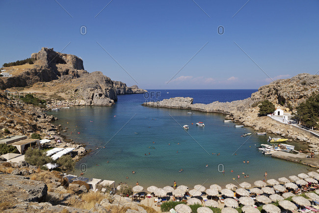 The Lindian Acropolis rises over St. Paul's Bay, a cove lined by sun shades, on a sunny day in Lindos on Rhodes, Dodecanese, Greek Islands, Greece, Europe
