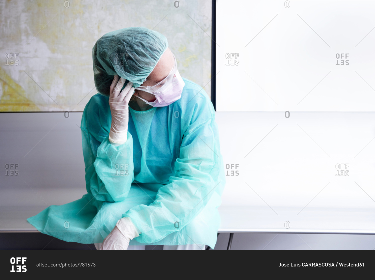 Sad doctor wearing protective workwear sitting against wall in hospital