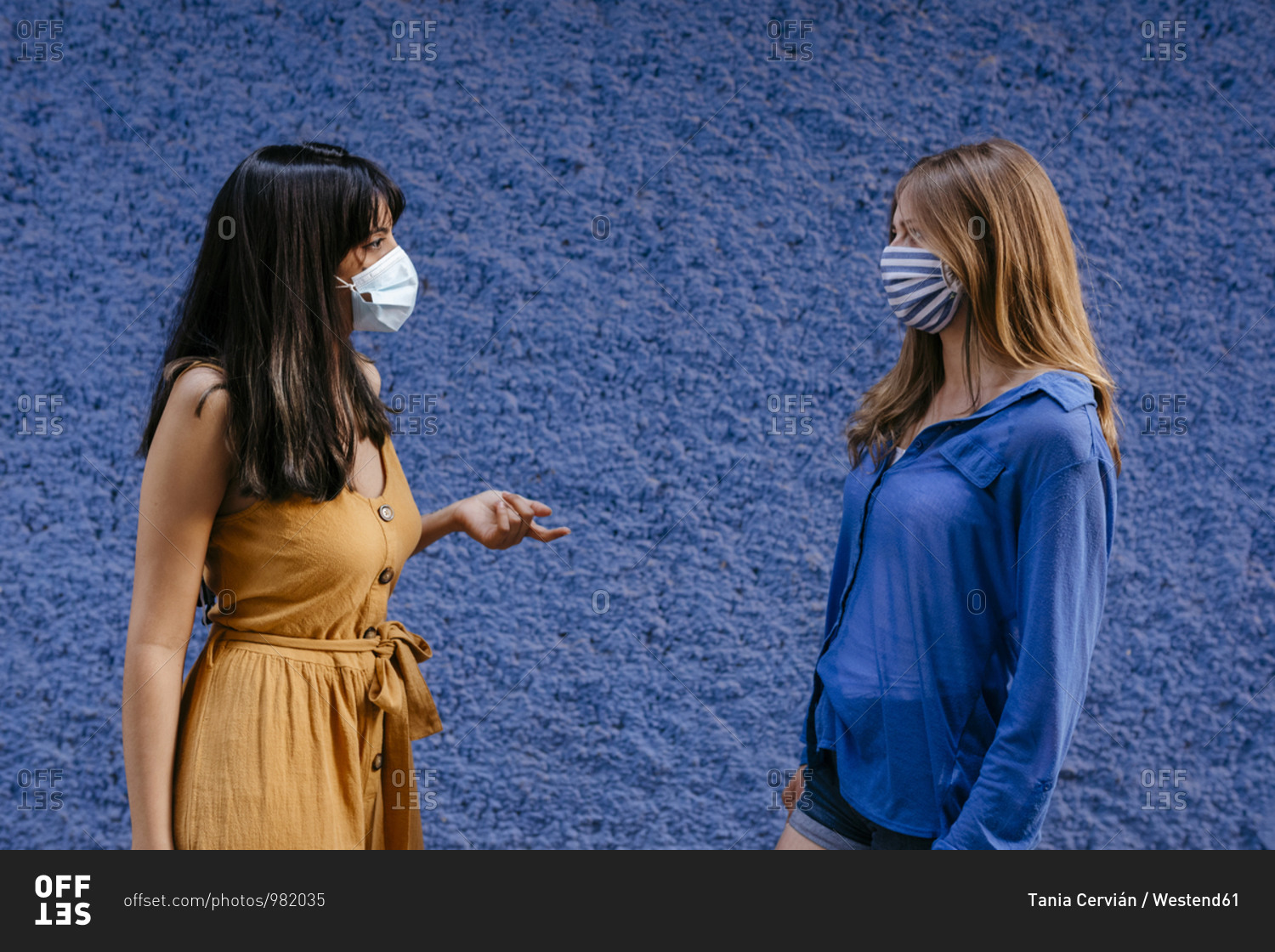 Female friends wearing masks talking while maintaining distance against blue wall in city
