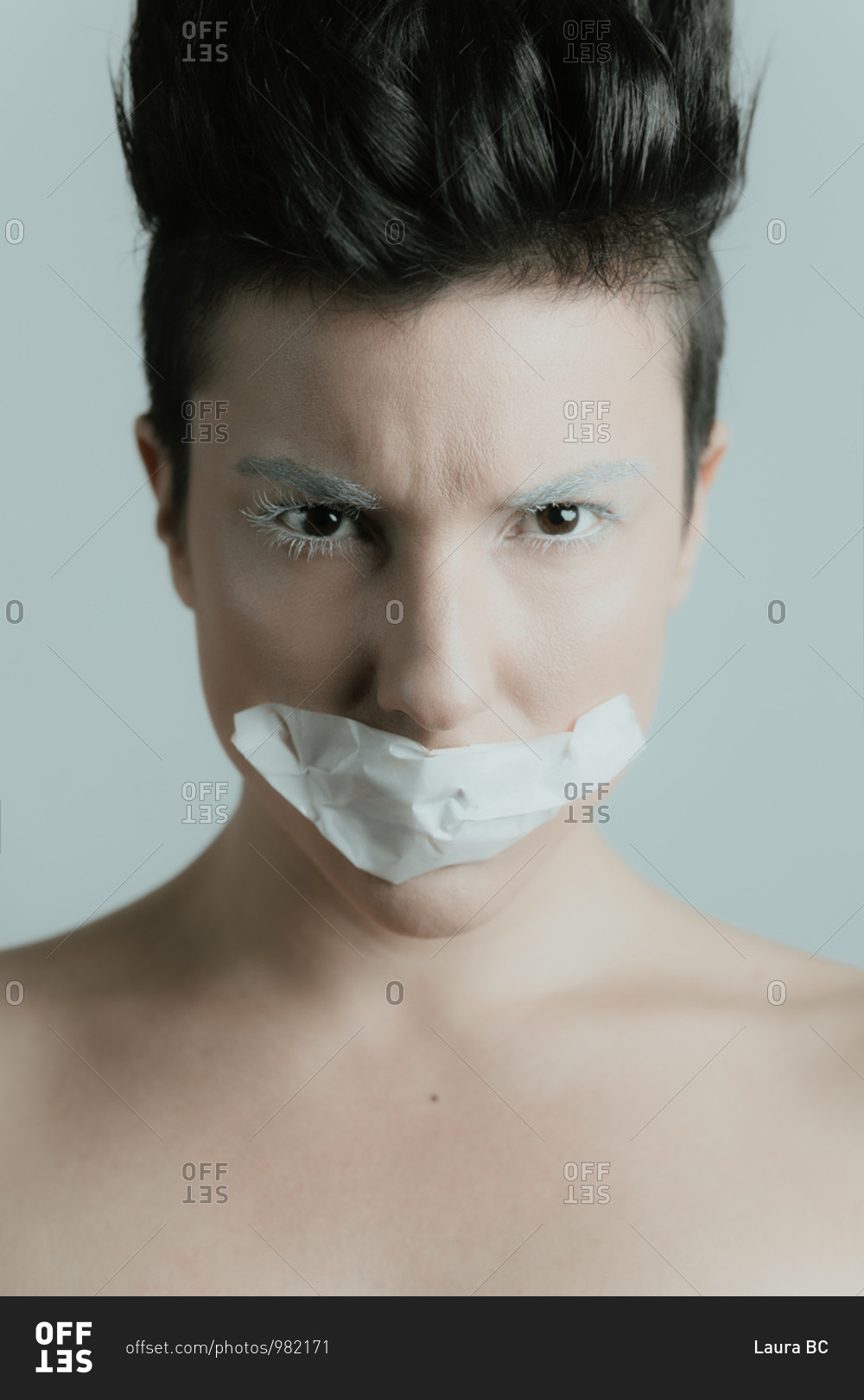 Creative portrait of a serious and attractive woman with short hair looking at camera wearing artistic white make up and tape over her mouth in studio