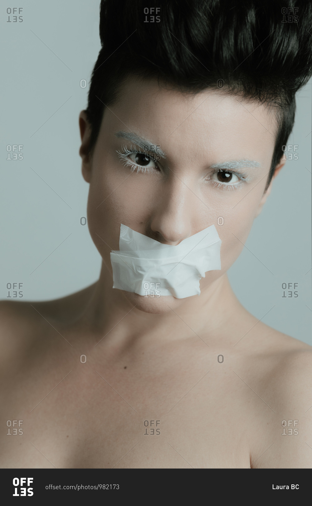 Creative portrait of an attractive woman with short hair looking at camera with her eyes closed wearing artistic white make up and tape over her mouth in studio