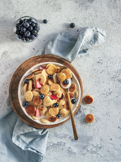 Trendy food - pancake cereal. Milk with cereal pancakes in bowl. Tiny cereal pancakes with berries in craft plate over gray cement background. Copy space