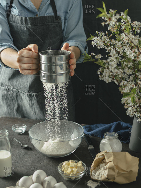 Young woman making cake. Female hands sifting flour from metal sieve in to glass mixing bowl on dark background. Food ingredients and kitchenware with flour, butter, milk and eggs at foreground and bouquet of blossoming cherry or pear branches. Photo seri