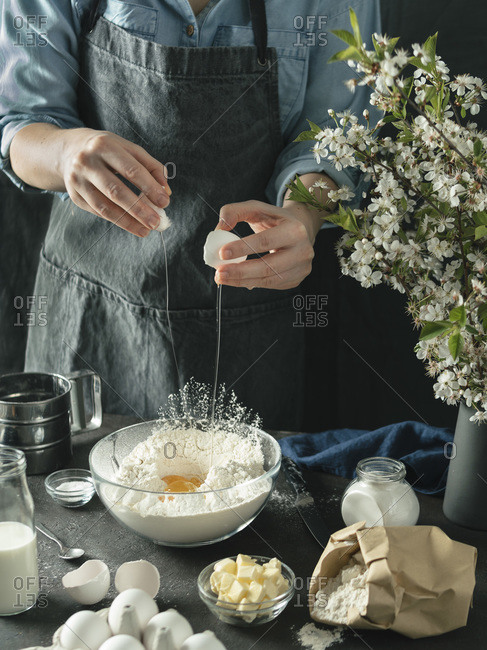 Young woman in blue shirt and gray apron making cake. Female hands break the egg in mixing boul with flour on dark background. Food ingredients and kitchenware with flour, milk, butter and eggs at foreground and bouquet of blossoming cherry or pear branches. Photo series.