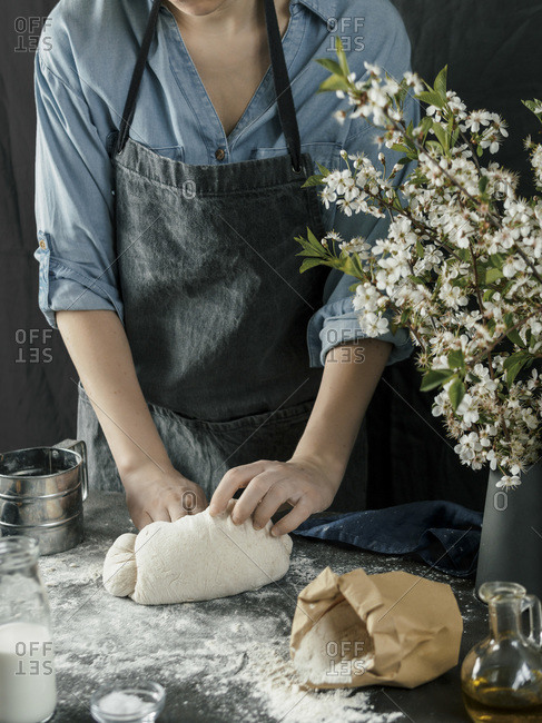 Young woman in blue shirt and gray apron making bread dough. Female hands kneading dough on dark kitchen. Black table with food ingredients - flour, milk, salt, olive oil and bouquet of blossoming cherry or pear branches. Home bread baking. Photo series.