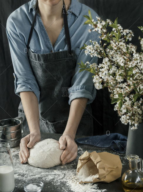 Young woman in blue shirt and gray apron making bread dough. Female hands kneading dough on dark kitchen. Black table with food ingredients - flour, milk, salt, olive oil and bouquet of blossoming cherry or pear branches. Home bread baking. Photo series.