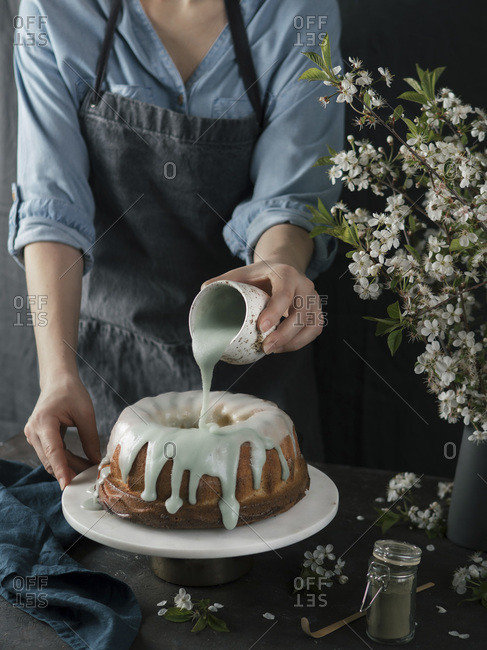 Young woman in blue shirt and gray apron pouring green matcha glaze on bundt cake on dark background. Bunt cake on marble cake stand and green tae matcha powder in glass jar and bouquet of blossoming cherry or pear branches. Photo series.