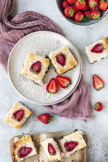 Two slices of coconut ice squares with chocolate bottom and strawberry topping on a white ceramic plate photographed on a light background. Fresh strawberries and a wooden serving board accompany.