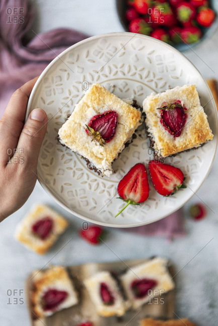 A hand holding a white plate with two slices of coconut ice squares with chocolate bottom and strawberry topping photographed on a light background. Fresh strawberries and a wooden serving board accompany.