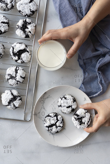 Woman holding a glass of milk and a flourless chocolate crinkle cookie on a plate, more cookies on a cooling rack accompany.