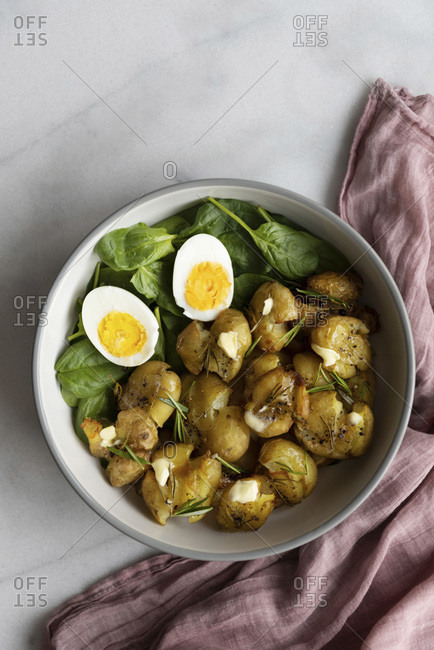 Smashed roasted potatoes in a salad bowl with baby spinach and hard boiled eggs photographed on a light marble background.
