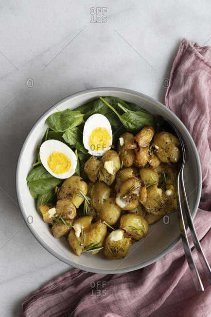 Smashed roasted potatoes in a salad bowl with baby spinach and hard boiled eggs with spoons photographed on a light marble background.