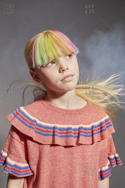 Portrait of girl with long blond hair and dyed fringe wearing pink frilly top, looking up, on grey background.