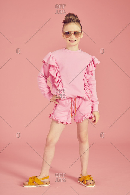 Portrait of brunette girl wearing pink frilly top, shorts and sunglasses on pink background.