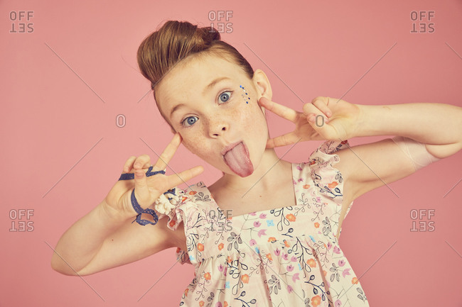 Portrait of brunette girl wearing frilly dress with floral pattern on pink background, sticking out tongue at camera.