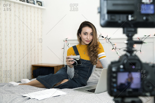 Young woman with long brown hair sitting on bed with camera and laptop, recording blog.