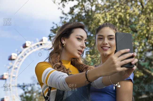 Two young women with long brown hair standing in a park near a Ferris wheel, taking selfie with mobile phone.