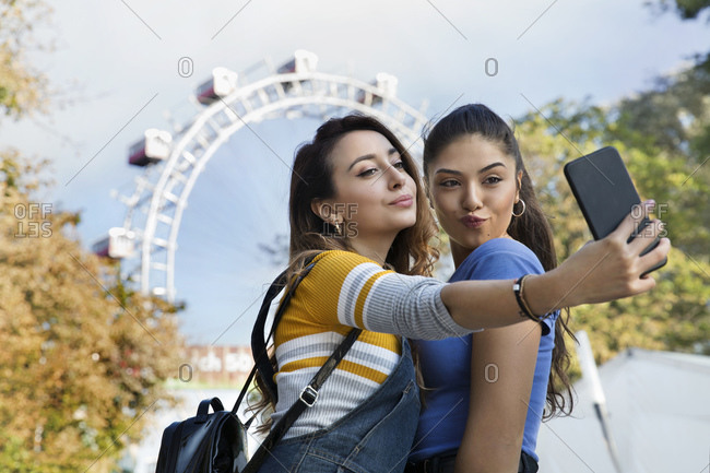 Two young women with long brown hair standing in a park near a Ferris wheel, taking selfie with mobile phone.