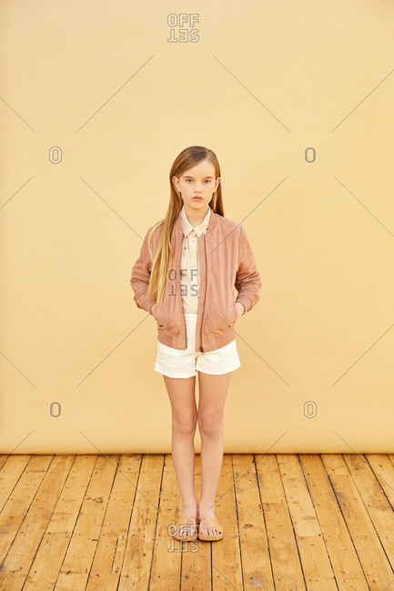 Portrait of girl with long blond hair wearing shorts, shirt and pink jacket, on pale yellow background.