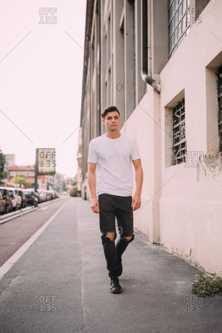 Portrait of young man with dark brown hair, wearing white T-shirt and ripped black jeans, walking down street.