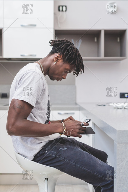 Young man with short dreadlocks sitting on bar stool in kitchen, using mobile phone.
