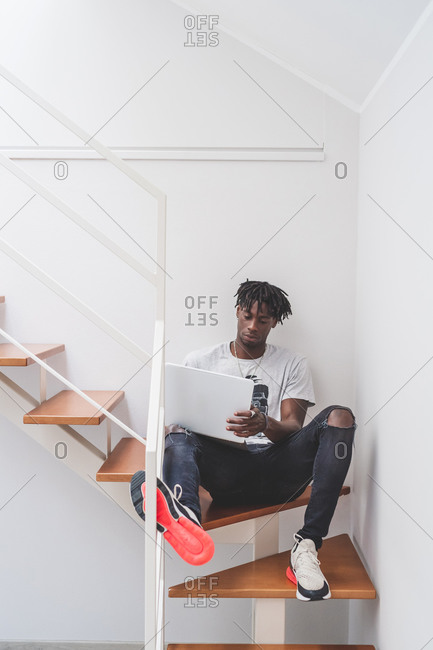 Young man with short dreadlocks sitting on staircase, typing on laptop.