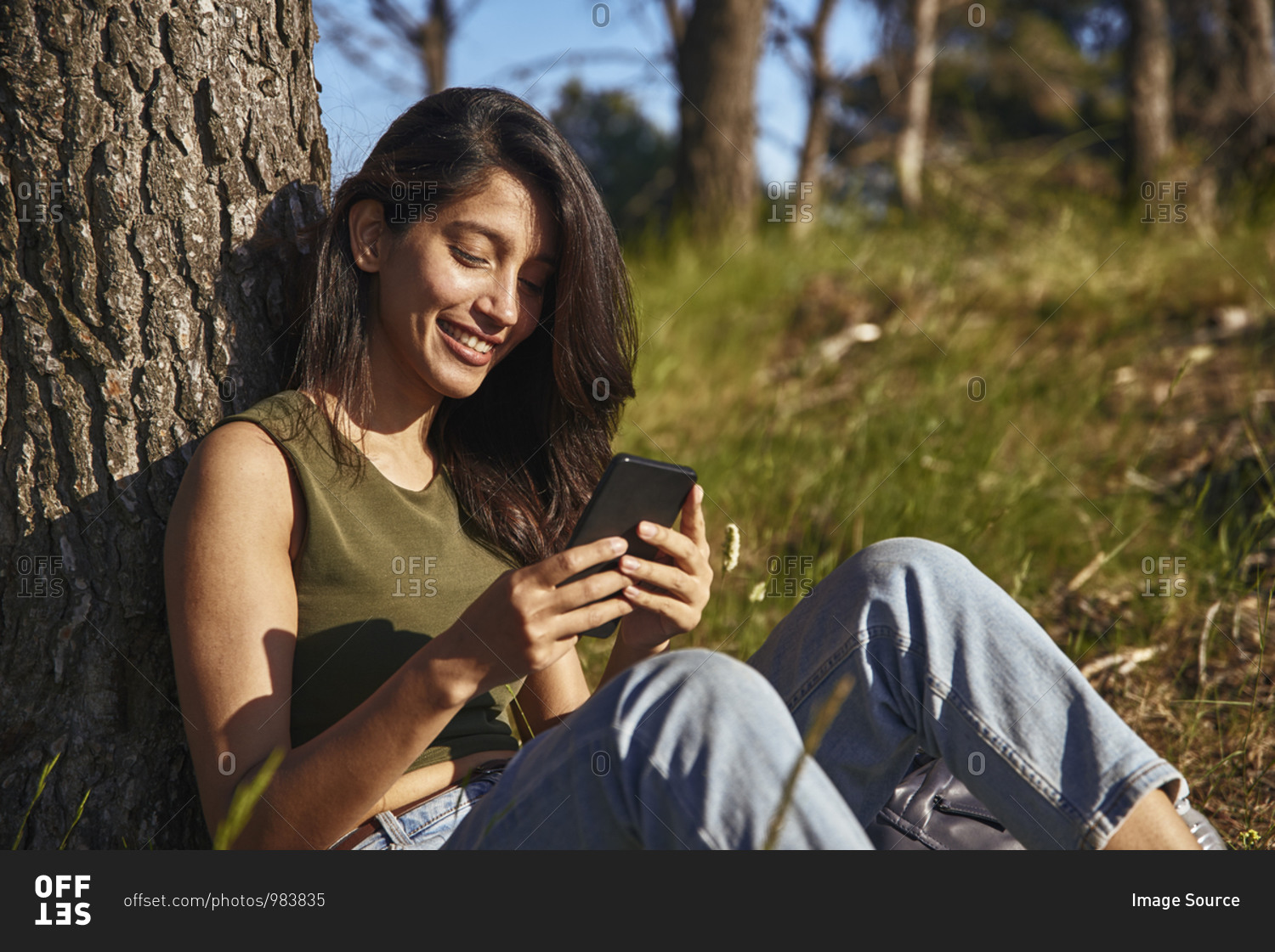 Portrait of young woman with long brown hair sitting under a tree in a forest, checking mobile phone.