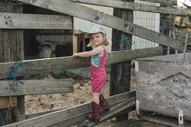 A young girl stands on a fence wearing a leotard and cowgirl boots.