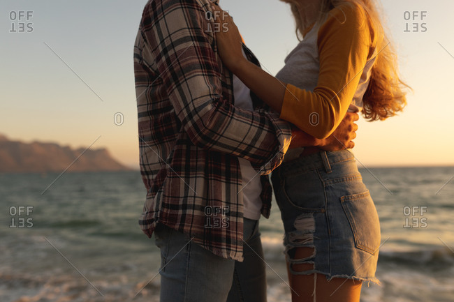 Mid section of a Caucasian couple standing on a promenade by the sea at sunset, facing each other and embracing. Romantic seaside holiday couple