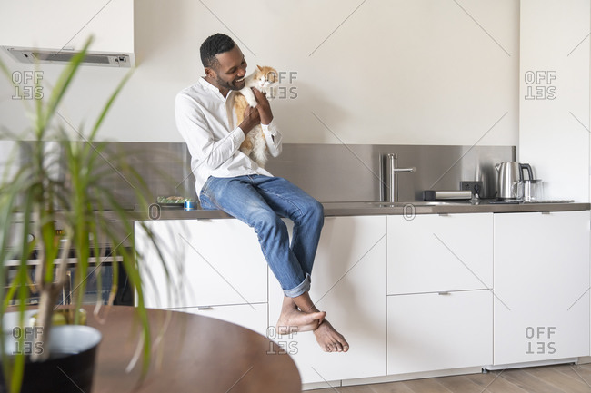 Portrait of smiling man sitting on kitchen counter at home cuddling his cat