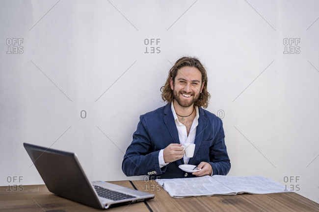 Happy young male entrepreneur sitting with espresso coffee and newspaper against white wall at cafe