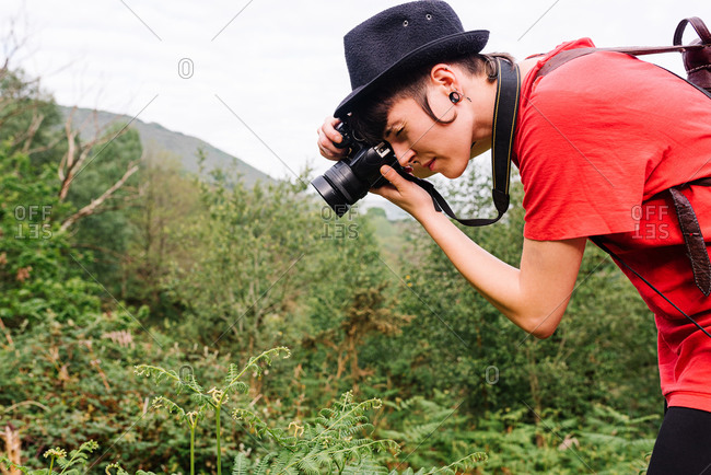 Side view of talented female photographer in trendy outfit taking photo of plant in field on photo camera