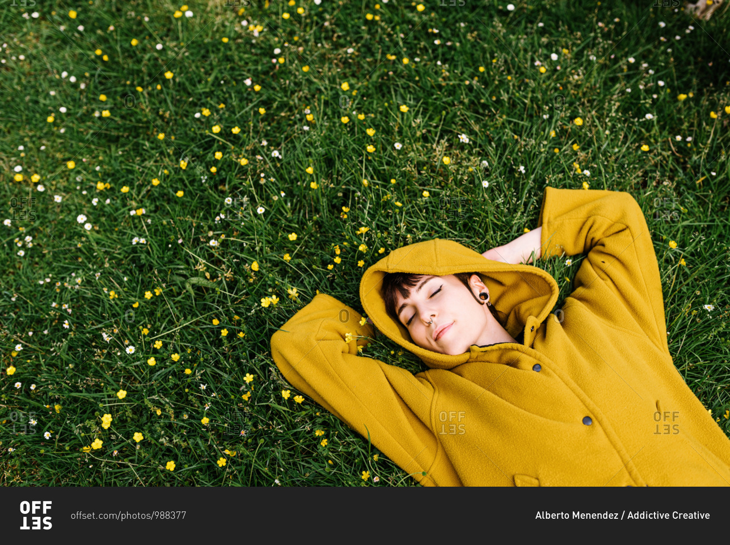 From above of content female with piercing resting with hands behind head on green lawn with flowers while enjoying summer