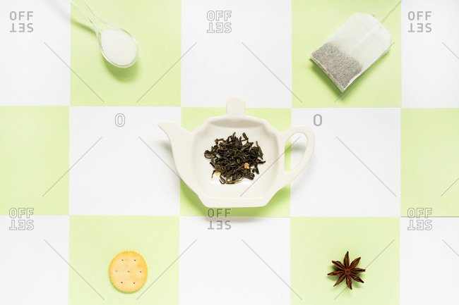 Top view of ceramic plate in form of teapot with green tea leaves near transparent tea bag with spoon of sugar and salty cracker with dry anise