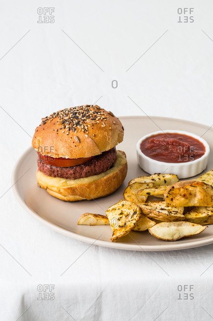 From above of delicious hamburger with meat cutlet and golden buns decorated with mix of sesame seeds near crunchy toasted potato slices with spices and salsa barbecue on ceramic plate