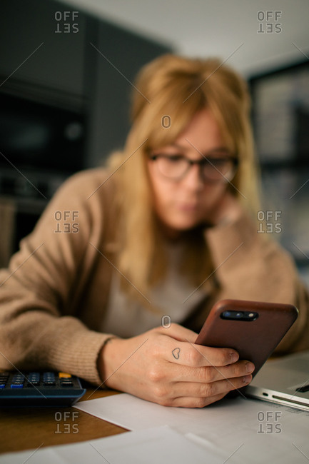 Side view of pensive female accountant sitting at table using on mobile phone while having break during remote work at home and laughing