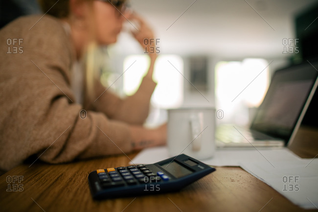 Focused blurred anonymous female bookkeeper sitting at table in kitchen and counting bills while doing paperwork using calculator