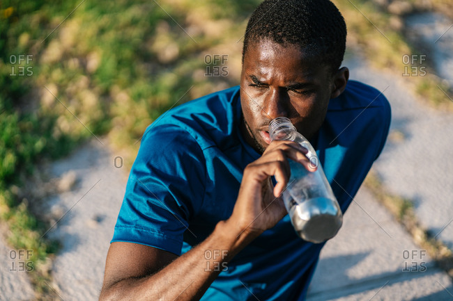 African athletic black man in sport clothes sitting down and drinking water from a bottle at sunset