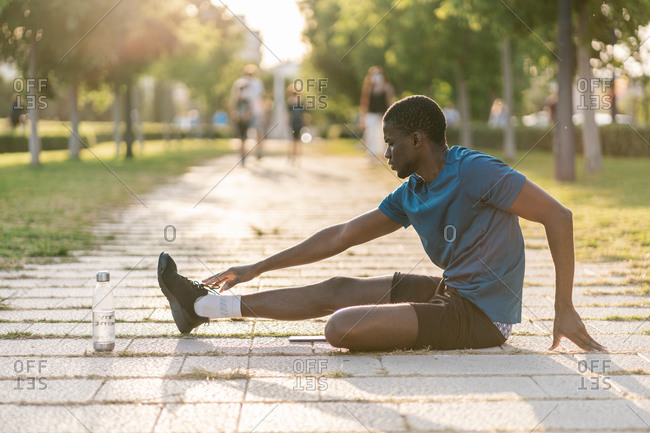 African athletic black man in sport clothes sitting on the floor and stretching in the park on a sunny day