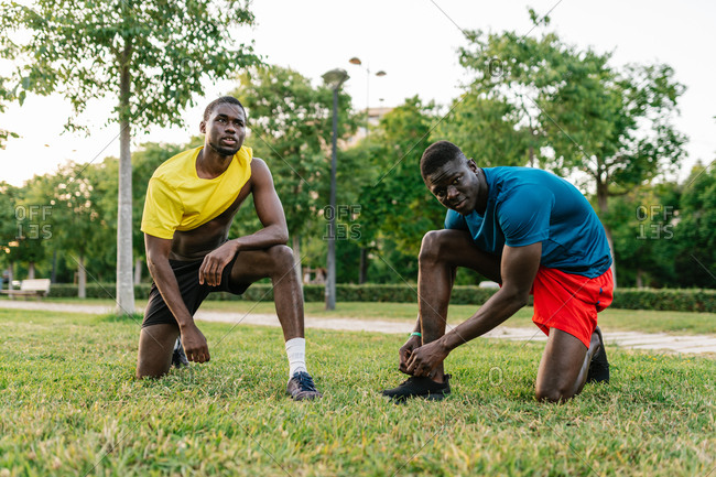 Two athletic black men wearing colorful sport clothes kneeling down resting after running in the park on a sunny day